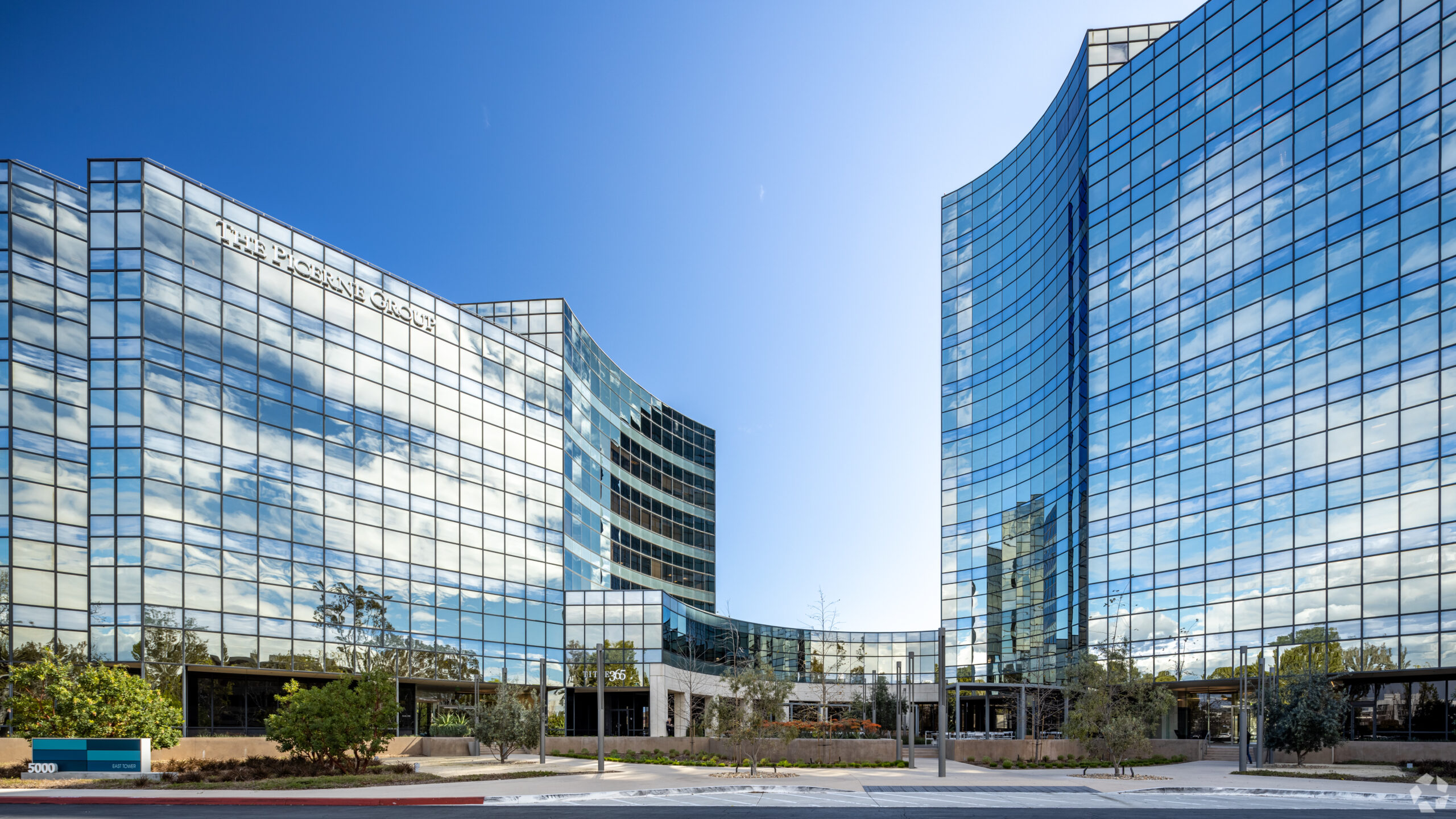 Two modern glass office buildings with mirrored windows reflect the blue sky and clouds, framing a central entrance area with landscaping in front. The sleek, professional facade is home to a leading digital marketing agency, its dynamic presence echoed in the contemporary architecture. The buildings rise several stories and are joined by a lower structure at the base.