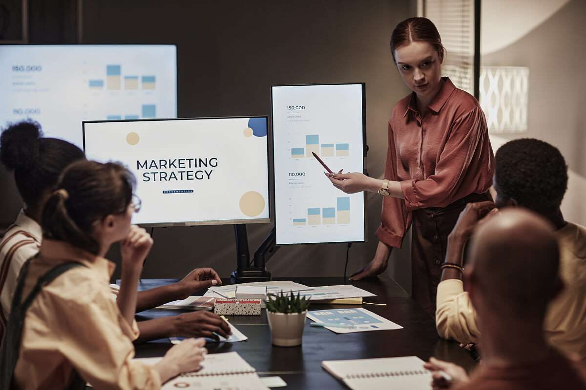 A woman stands and presents marketing data on screens to three seated colleagues in a dimly lit office during a digital marketing agency meeting.