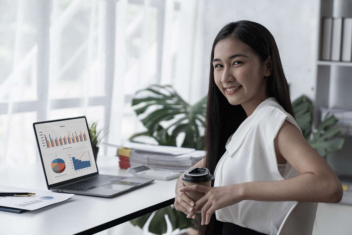 A woman sits at a desk, smiling while holding a coffee cup. Beside her, a laptop displaying charts and graphs—perhaps showcasing analytics for a digital marketing agency—is open, with paperwork stacked neatly on the desk. Large windows and lush plants are visible in the background, creating an inviting workspace.