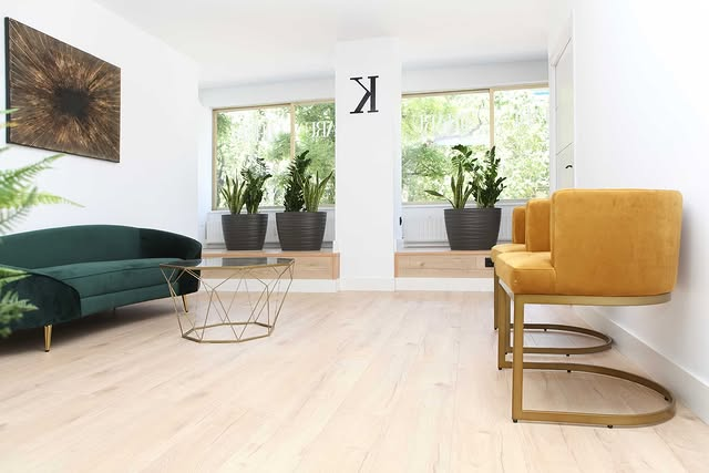 Modern living room with a green velvet sofa, two mustard yellow chairs, a geometric glass coffee table, large potted plants by the window, and gold abstract wall art. Light wood flooring and white walls create a bright space—perfect for brainstorming sessions or creative meetings if you’re running a digital marketing agency from home.