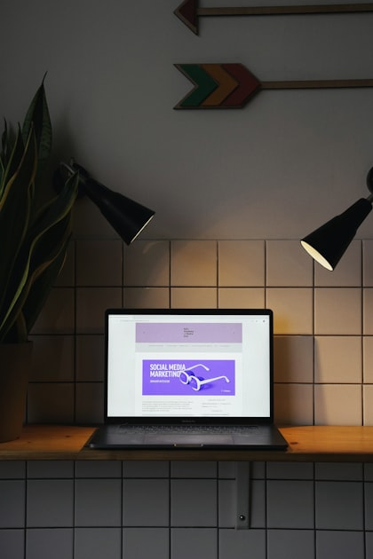 A laptop on a wooden shelf displays a purple Social Media Marketing page, flanked by two black lamps. A plant and colorful arrows decorate the tiled wall behind, reflecting the creative flair of a full service marketing agency.