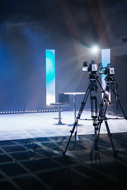 Two video cameras on tripods face an empty modern stool and table on a brightly lit stage, with blue and white vertical lights in the background—perfect for filming content marketing packages or showcasing the expertise of a leading SEO agency.