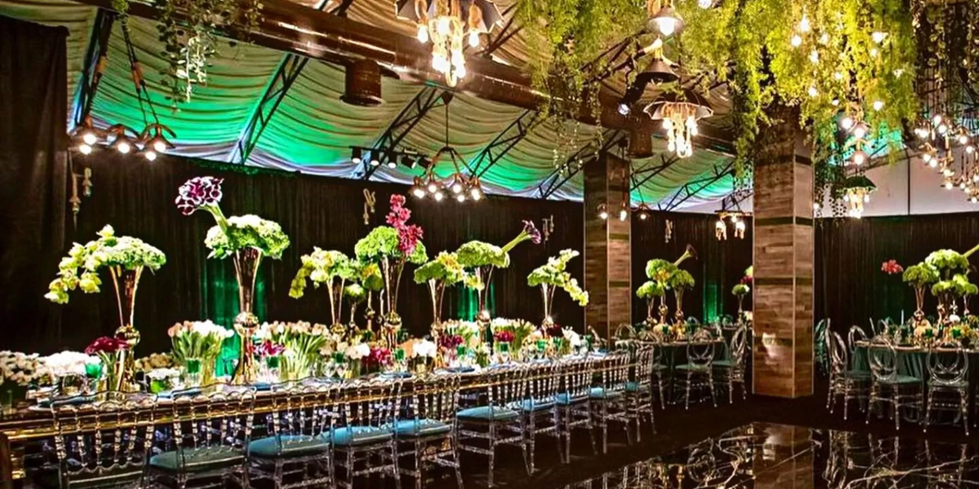A luxurious banquet hall with long tables decorated with tall green floral centerpieces, elegant place settings, clear chairs, hanging greenery, chandeliers, and green accent lighting—perfect for hosting an unforgettable event or celebrating the achievements of your digital marketing agency in style.
