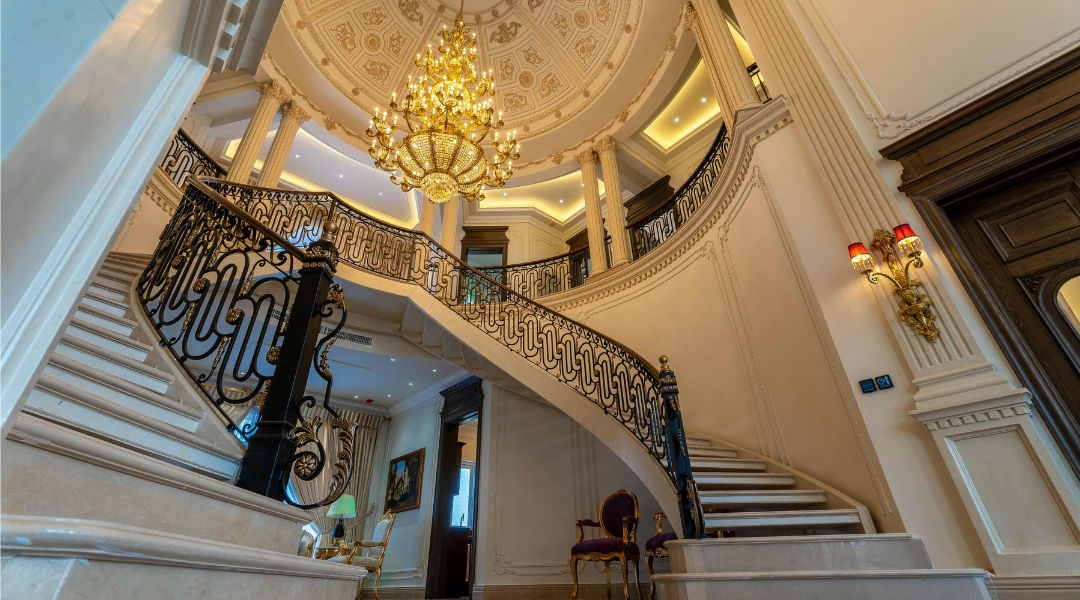 Grand staircase with ornate black railings, marble steps, a large chandelier, domed ceiling, classic columns, elegant wall sconces, and luxurious decor create a lavish, upscale interior—an impressive space well-suited for an innovative digital marketing agency.