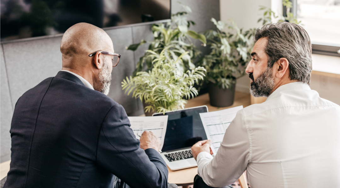 Two men sitting at a table discussing paperwork, with a laptop open in front of them and plants in the background, strategizing for their digital marketing agency.